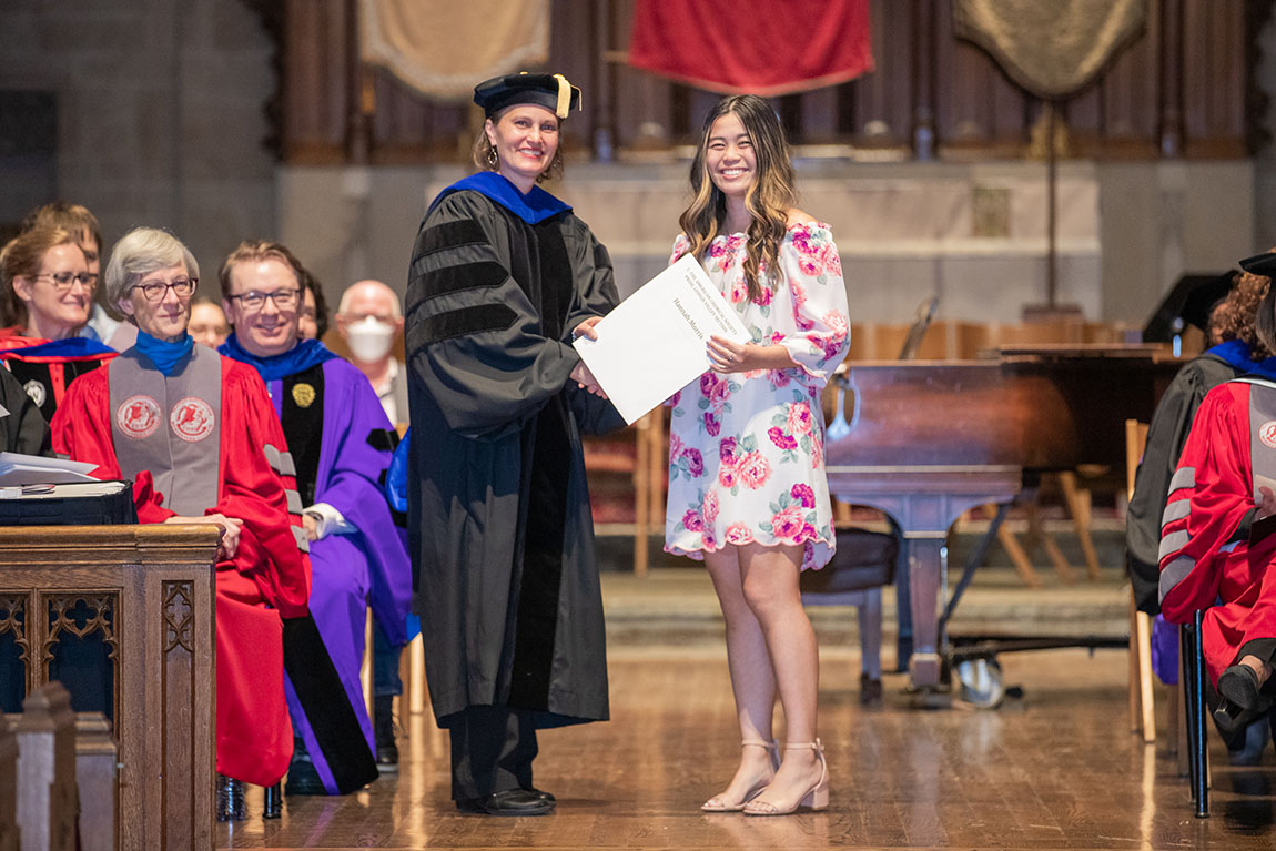 A woman in black academic robes and hat shakes hands with a college student with long hair wearing a flowery dress